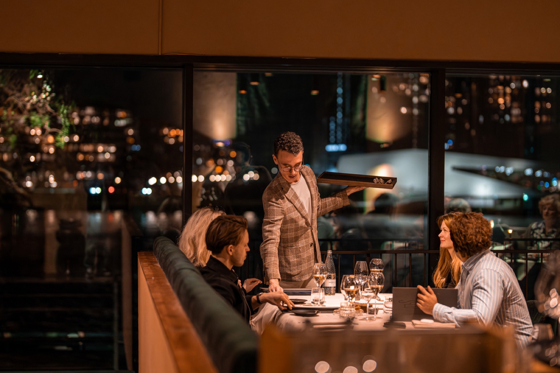 Gasten in gesprek aan tafel in MOS, met de lichtjes van Amsterdam achter het raam